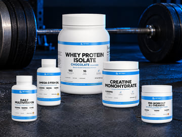 The Advanced stack on a dark gym floor beside a loaded barbell: Daily Multivitamin, Omega-3 Fish Oil, Whey Protein Isolate (chocolate), Creatine Monohydrate and Pre-Workout & L-Theanine (fruit punch).